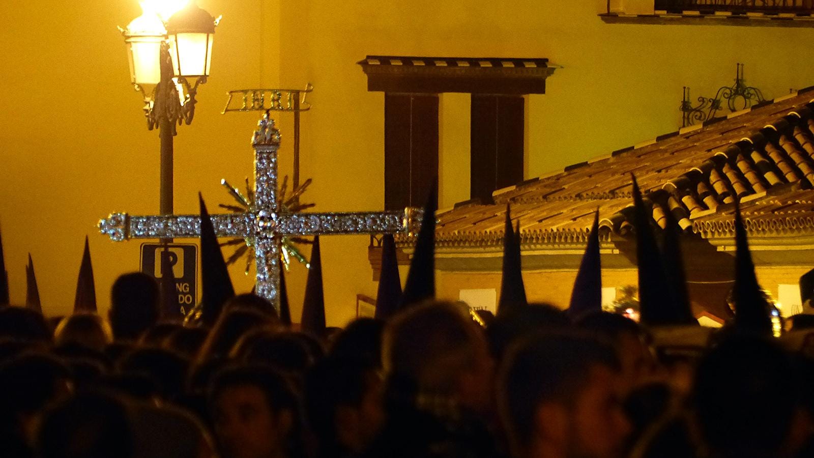 Cruz procesional y nazarenos entre una multitud durante la Semana Santa de Sevilla.