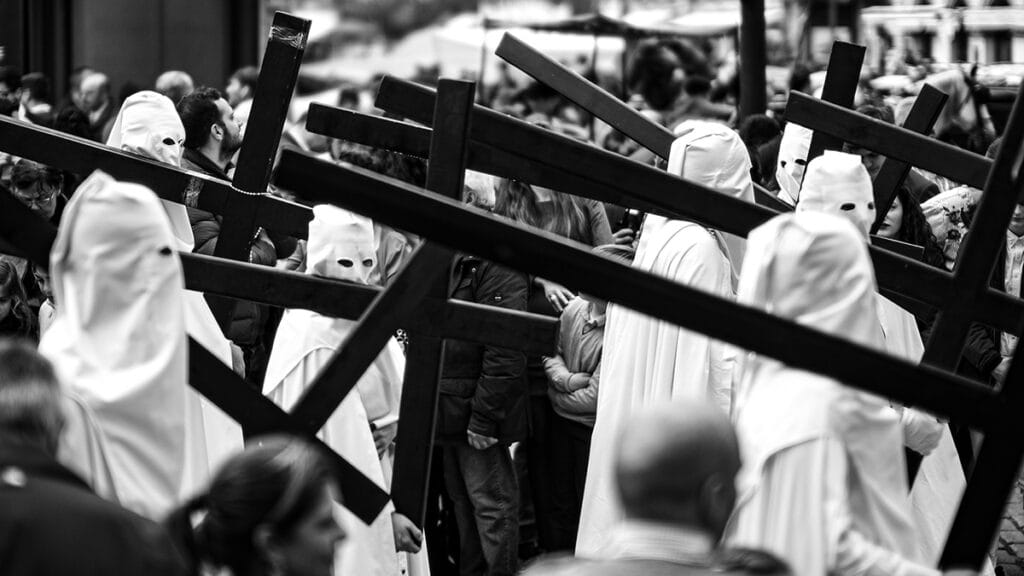 Penitentes portando cruces entre el público durante la Semana Santa de Sevilla, en una fotografía en blanco y negro.