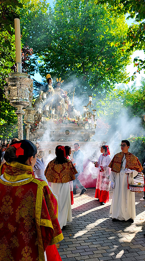 Acólitos con vestiduras litúrgicas caminando entre árboles y una nube de incienso durante el traslado matinal.