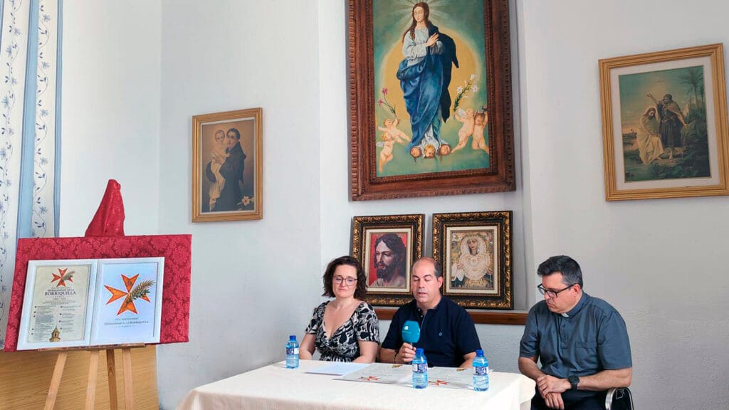 La alcaldesa de Padul, Celia Villena, el hermano mayor de la Borriquilla, Andrés Rejón, y el párroco, Carlos José Fernández, sentados a una mesa presidiendo la presentación del XXV Aniversario en los Salones Parroquiales.