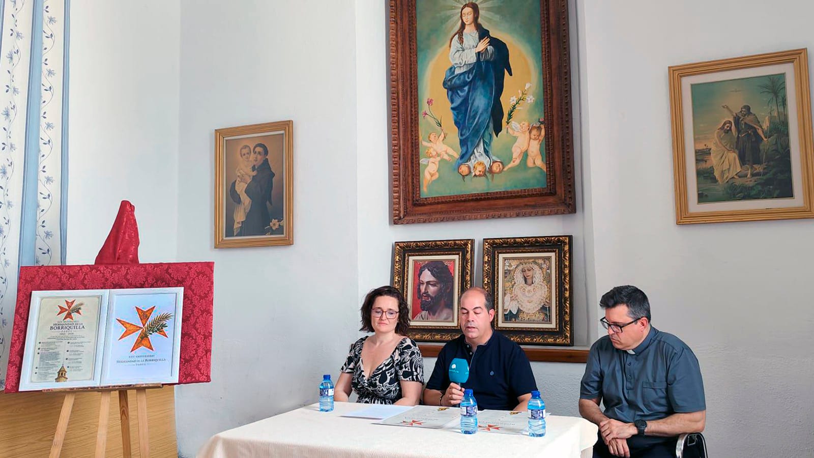 La alcaldesa de Padul, Celia Villena, el hermano mayor de la Borriquilla, Andrés Rejón, y el párroco, Carlos José Fernández, sentados a una mesa presidiendo la presentación del XXV Aniversario en los Salones Parroquiales.