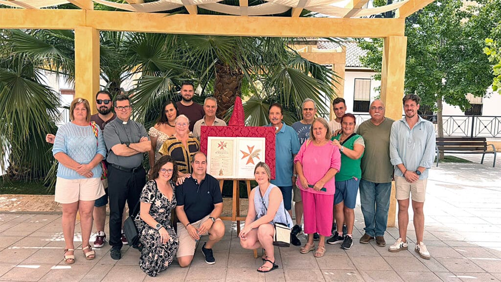 Foto de grupo con miembros de la Hermandad de la Borriquilla y asistentes posando al aire libre en un patio alrededor del cartel y el logotipo del XXV Aniversario expuestos en un caballete.