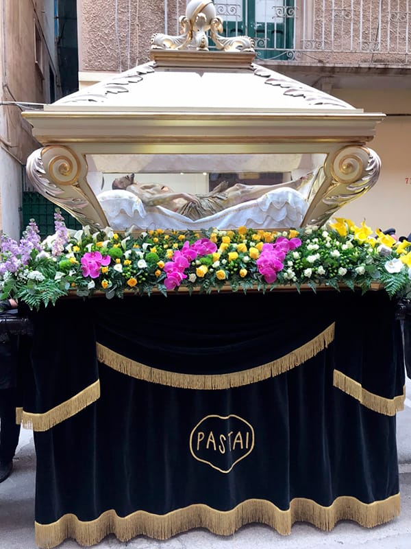 Jesús yacente en urna procesional, acompañado de flores, en la Semana Santa de Trapani.