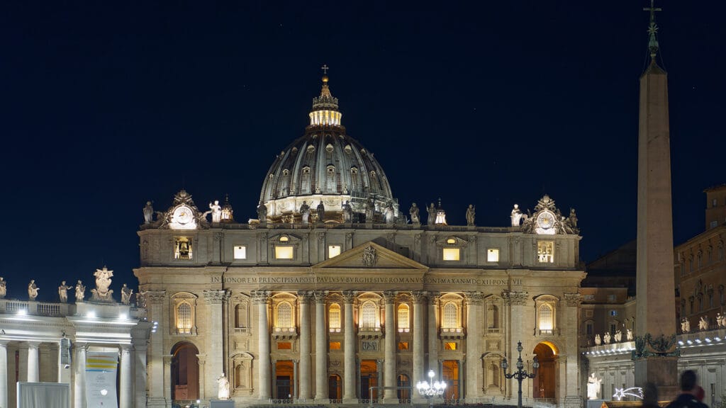 Fachada de la Basílica de San Pedro iluminada de noche, con la plaza y el obelisco en primer plano.