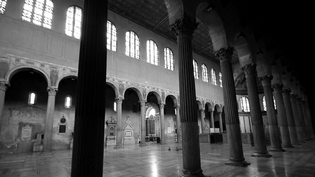 Interior de la basílica de Santa Sabina en Roma, con largas hileras de columnas y nave central iluminada.