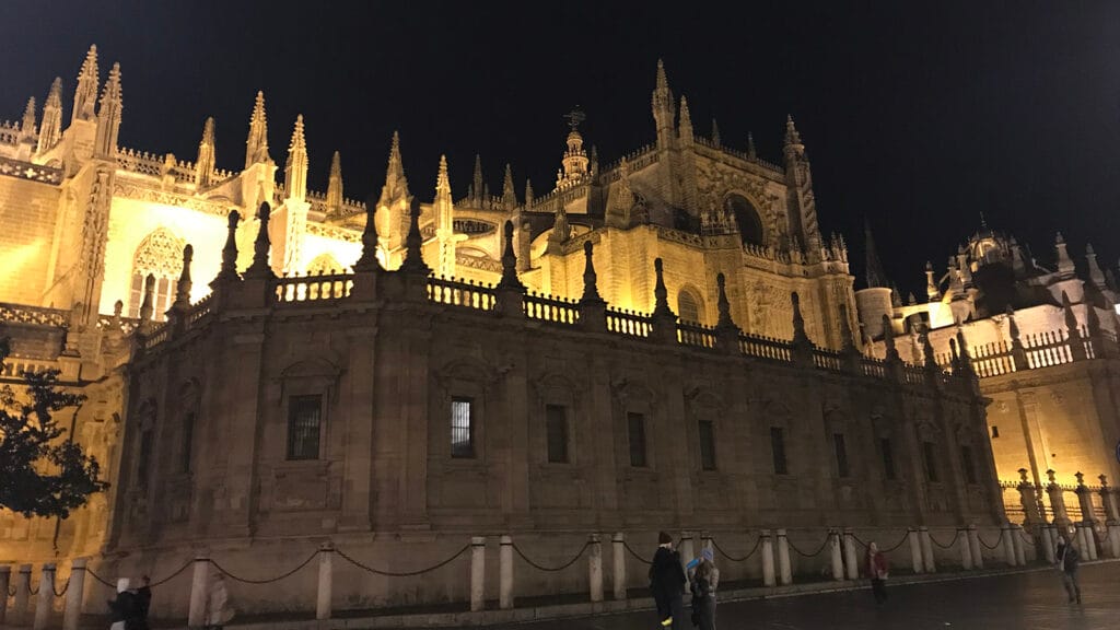 Catedral de Sevilla iluminada de noche, con la Giralda destacando sobre el cielo oscuro y la fachada bañada por luz artificial.