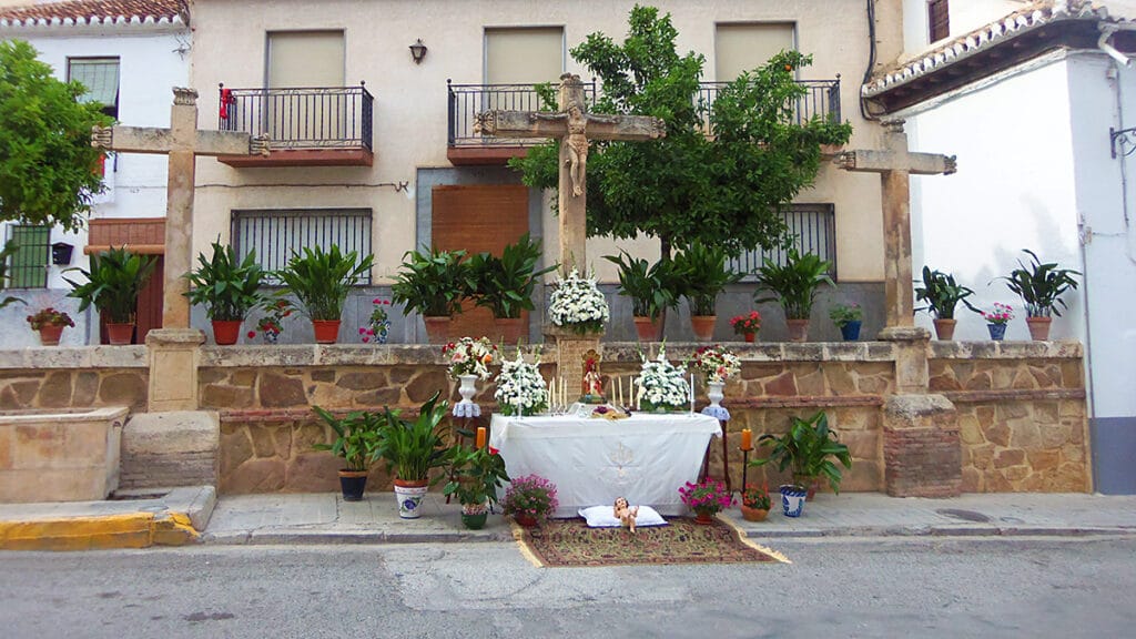 Altar efímero en la calle Real de Padul: tres cruces de piedra al fondo y mesa cubierta una tela blanca bordada, velas y arreglos florales, preparado para el paso de la procesión.