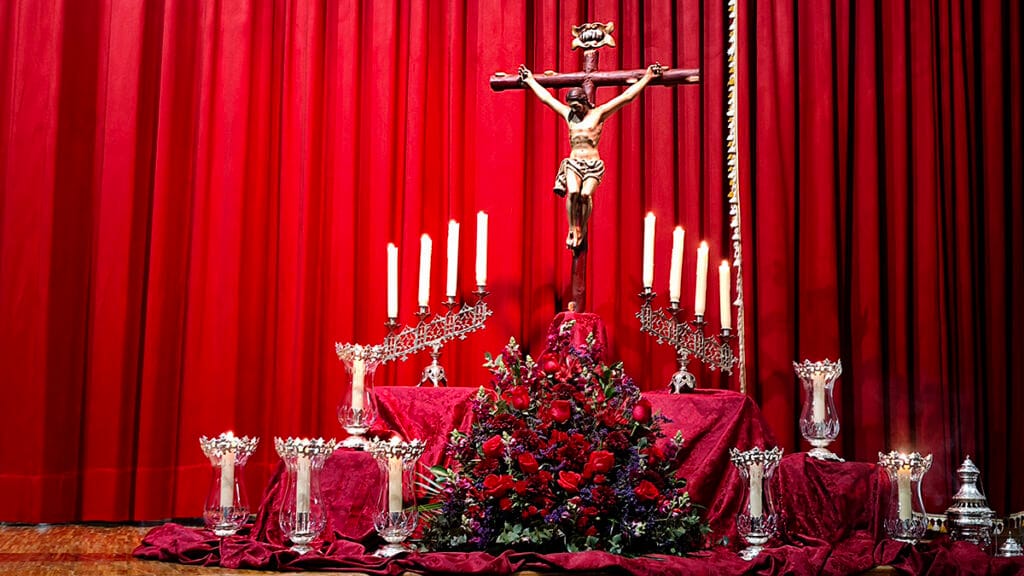 Decoración del escenario del pregón con crucificado, candelabros y centro floral en tonos rojos.