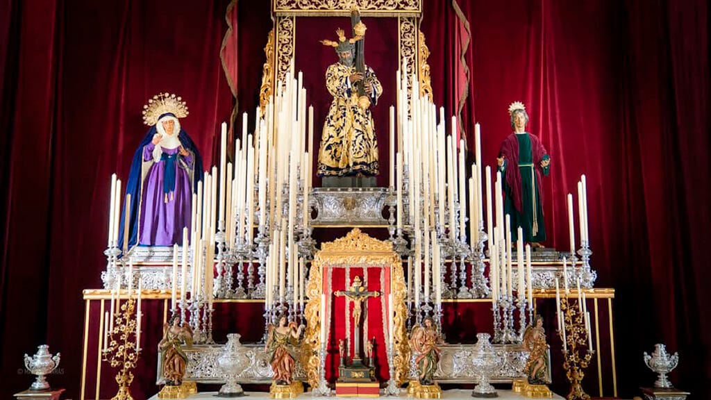 Altar de cultos de la Hermandad de Los Gitanos de Sevilla. En el centro, la imagen del Señor de la Salud con túnica bordada y cruz al hombro sobre un pedestal de plata. A la izquierda, la Virgen de las Angustias con manto azul y saya morada; a la derecha, San Juan Evangelista con túnica verde y mantolín rojo. El conjunto está decorado con decenas de cirios blancos simétricos y un fondo de cortinajes de terciopelo burdeos.