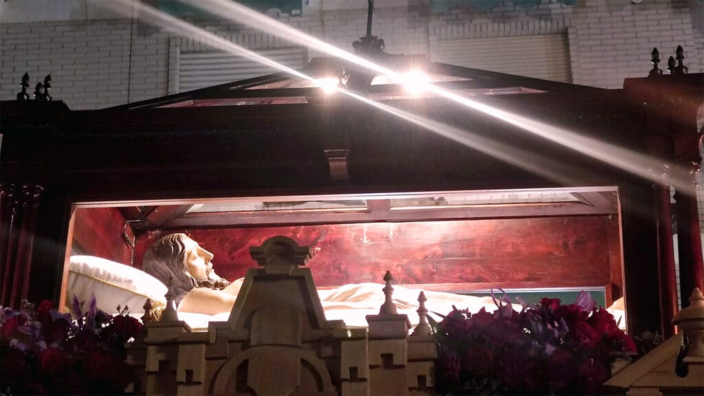Cristo yacente en urna acristalada del Santo Sepulcro durante la noche del Viernes Santo en Padul, iluminado por focos y rodeado de flores.